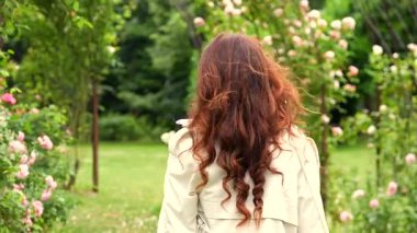 Uzun dalgalı saçlı, açık renk trençkot giyen, baharda çiçek açan bir gül bahçesinde yürüyen bir kadının dikiz aynasından. Çiçeklerle çevrili ve yemyeşil, yumuşak doğal ışıkla..