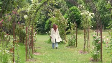 Bahar ya da yaz aylarında sakin yeşil bir bahçede çiçek açan güllerle kaplı metal kemerlerin altında yürüyen bir kadının arka görüntüsü. Yumuşak doğal ışık ve sakin mevsimsel atmosfer.