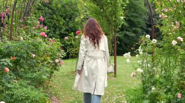 Açık renk montlu bir kadının, bir bahar günü yeşilliklerle çevrili sessiz bir bahçedeki çiçek açan gül çalılarının arasında gezinişi. Yumuşak doğal ışıkla huzurlu bir çiçek ortamı..