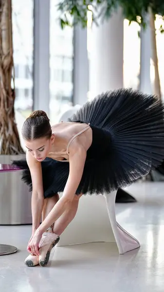 Siyah etekli balerin sivri uçlu ayakkabılarını ayarlamak için öne doğru eğilir, parlak bir dans stüdyosunda parlak beyaz bir sandalyeye oturur ve ışık saçar.