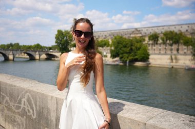 Beyaz elbiseli neşeli bir kadın Paris 'te Seine Nehri' nin kenarında duruyor, gülümsüyor ve güneşli bir yaz gününün tadını çıkarıyor. 