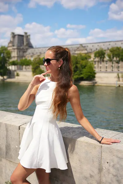 Beyaz elbiseli ve güneş gözlüklü kendinden emin bir kadın Paris 'te Seine Nehri yakınlarında poz veriyor. Güneşli bir günde Louvre Sarayı' nın arka planında ve neşeli bir ifadeyle. 