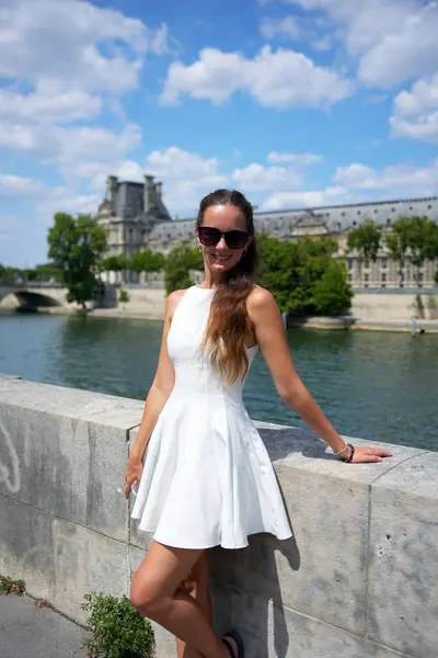 Güneşli bir yaz gününde Paris 'teki Seine Nehri' nin kenarında duran beyaz elbiseli, kendine güvenen, arka planda klasik mimarisi olan bir kadın. 