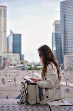 Bir kadın, arka planda gökdelenleri ve şehir hayatı olan La Defense Paris 'te otururken kitap okuyor. 
