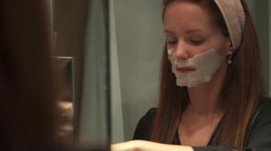 Evdeki cilt bakımı ritüelinin bir parçası olarak banyo aynasının önüne maske takmaya hazırlanan genç bir kadın. 