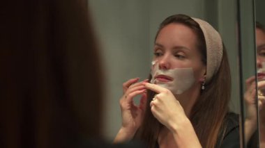 Genç bir kadın akşam güzelliği sırasında aynada kendine bakarken cildine bir maske takıyor. 