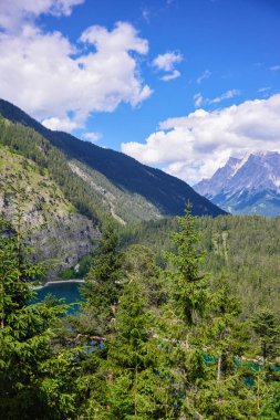 Yoğun yeşil ormanlarla çevrili dağ zirveleri ve parlak yaz gökyüzünün altında kabarık bulutlarla çevrili alp gölü manzarası..