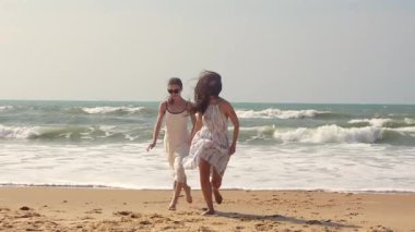 Kumsalda el ele yürüyen iki kadın dalgalı saçları ve açık renk elbiseleriyle