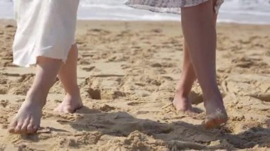 Beyaz yaz elbiseleriyle, kumsalda çıplak ayakla yürüyen iki kadının yakın çekimi.