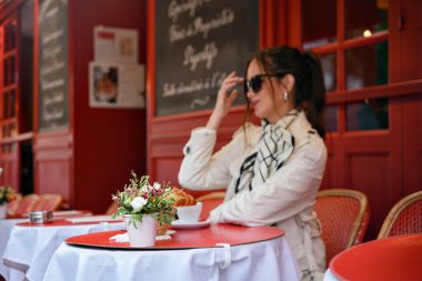 Modayı takip eden bayan kırmızı teras masasında otururken gözlüklerine dokunuyor.