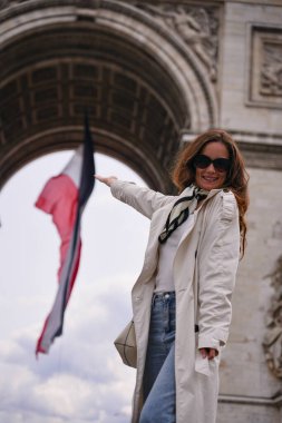 Açık renk trençkotlu, gururlu bir kadın Paris 'te Zafer Takı yakınlarında Fransız bayrağı taşıyor. Ulusal gurur ve tarzı simgeliyor. 