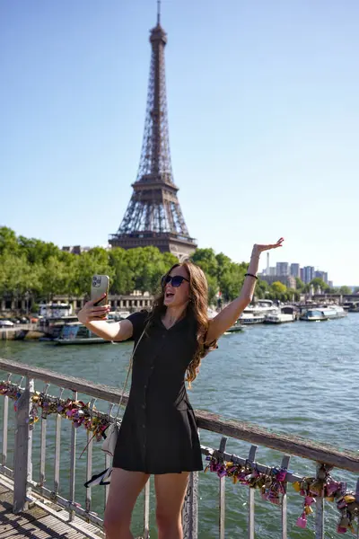 Genç kadın Paris 'te Eyfel Kulesi ile birlikte Seine Nehri üzerindeki köprüde selfie çekiyor.