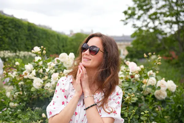 Çiçekli elbiseli ve güneş gözlüklü mutlu kadın beyaz gül çalılarının yanında duruyor.