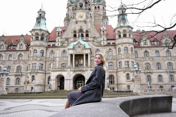 Tarihi belediye binasının önündeki taş duvarda oturan ceketli genç kadın.