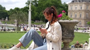 Lüksemburg Bahçeleri 'nde akıllı telefon kullanan trençkotlu şık bir kadın Paris' te. 