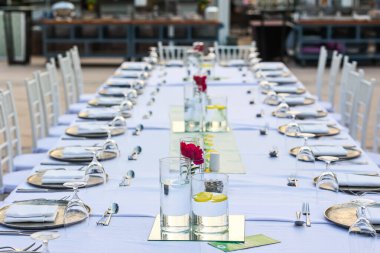 Etkinlik için şık bir açık hava ziyafet masası, beyaz sandalyeli güzel bir açık hava ziyafet masası, güzel yemek takımları, çiçekli süsler ve cam süsler, resmi bir etkinlik için ayarlanmış..  