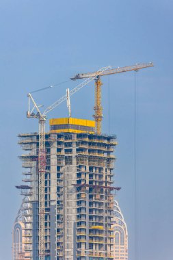 Üzerinde vinçler olan canlı bir şehir manzarasında inşaat halindeki yüksek katlı bir gökdelen. Modern mimari yapı tamamlanmış kuleler ve ön planda yemyeşil alanlar ile çevrilidir..