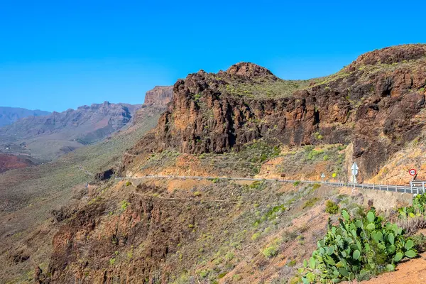 Kurak kanyon arazisi ve mavi gökyüzünün altındaki seyrek bitki örtüsüyle çevrili dik kayalık uçurumlar boyunca uzanan sarmal dağ yolu.. 