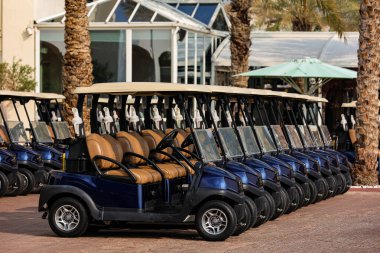 Golf arabaları ile lüks bir golf tesisi. Yüksek kaliteli fotoğraf, kemerli balkonları ve palmiye ağaçları olan şık tatil köyü binası, önünde uzun sıra modern golf arabaları var.. 