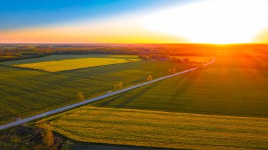 Uçsuz bucaksız yeşil tarlaların insansız hava aracı görüntüsü ve sıcak günbatımı ışığıyla aydınlatılan bir köy yolu huzurlu bir kırsal alan yaratıyor.. 