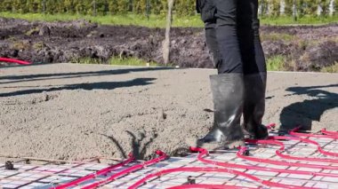 Video of a construction worker in rubber boots pouring concrete over red underfloor heating pipes during a foundation installation. High quality 4k footage