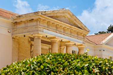 Bu resim, sütunlarla süslenmiş, yemyeşil ve parlak mavi gökyüzünün altında yuva yapmış, sükunet ve nostalji hissi uyandıran klasik tapınak benzeri bir yapı yakalıyor..