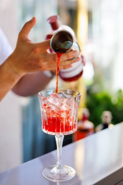 Barmen bardağa kırmızı kokteyl dökerken, barmenin bardağa buz küpleriyle parlak kırmızı kokteyl dökerken, bardakları karıştırırken, içkileri tazelerken, bardakları tazelerken.