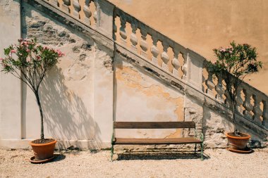 Tahta bir bank, tarihi bir binada güneşli bir terasa çıkan taş basamakların yanında saksılarla çevrilidir.