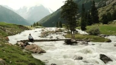Huzurlu ve huzurlu bir manzara, nefes kesen dağlık bir manzaranın ortasında akan bir nehrin kenarında oturan bir adamı anlatıyor.