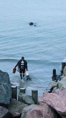 Yalnız bir figür kayalık bir sahilde duruyor, düşünceli bir şekilde sakin sulara bakıyor, muhtemelen balık tutuyor ya da keşfediyor.