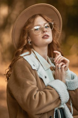 Sonbahar parkında poz veren şapkalı ve gözlüklü şık bir kız.