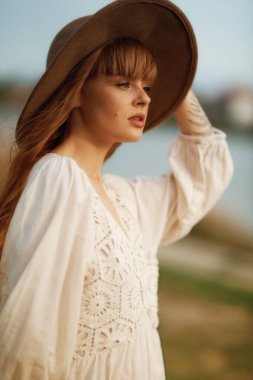 Beyaz gömlekli ve şapkalı genç kadın doğada poz veriyor.
