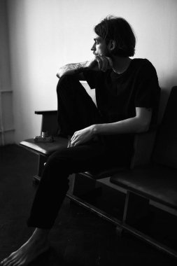 Siyah gömlekli ve kot pantolonlu bir adam sandalyeye oturur ve elinde bir akıllı telefon tutar.