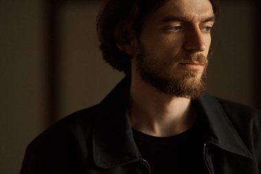 Siyah tişörtlü ve kot pantolonlu genç sakallı adam karanlık odada ciddi ciddi bakıyor..