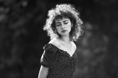 Genç, güzel esmer, kıvırcık saçlı, siyah elbiseli, yeşil arka planda poz veren kadın.