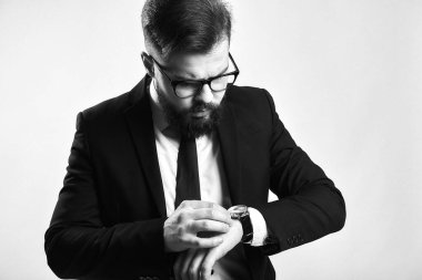 Ciddi yüzlü sakallı bir iş adamı, siyah takım elbiseli, kravatlı, gözlüklü, gri arka planda kol saati olan vahşi beyaz bir erkek..
