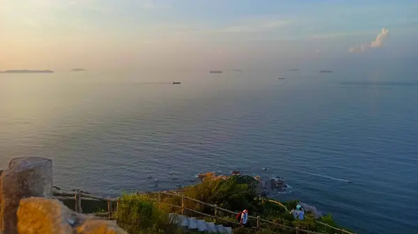 Sahil yolundaki yürüyüşçüler uzak gemilerle sakin bir denize bakıyor.