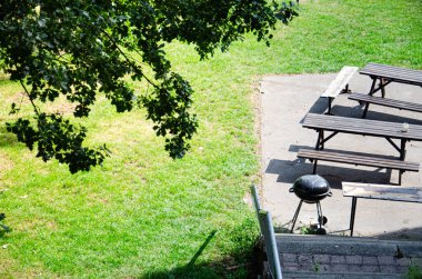 Barbekü ızgarası, ahşap bankları ve yeşil çimenlikteki büyük bir meşe ağacının yanındaki masaları olan bir oturma alanı. Güneşli bir gün, manzaralı, insan yok..