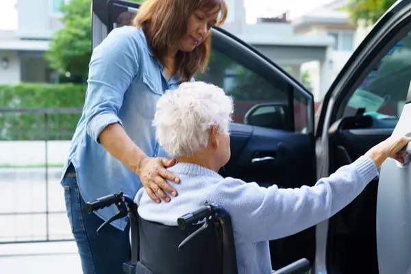 Bakıcı, Asyalı yaşlı kadın maluliyet hastasına arabaya binmek için tekerlekli sandalye kullanmasında yardım ediyor..