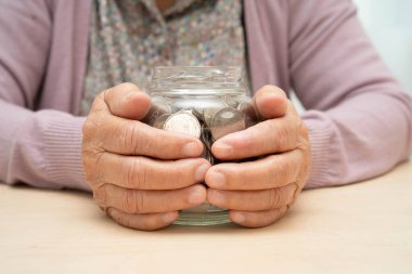 Retired elderly woman putting coins money in piggy bank and worry about monthly expenses and treatment fee payment.