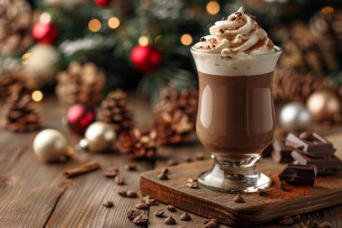 Bir bardak krem şantili ve tarçın çubuklu sıcak çikolata, yıldız anasonu ve çikolata parçacıklarıyla çevrili, ahşap bir masanın üzerinde oturur. Sıcak içecek, kırmızı ve kırmızı süslemeli bir Noel ağacının bulanık arka planının önünde duruyor.