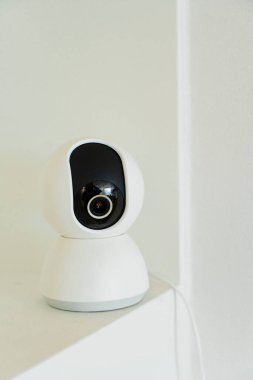 Beyaz, küresel bir güvenlik kamerası beyaz bir duvara karşı bir rafta duruyor ve ev izleme ve koruma sunuyor..