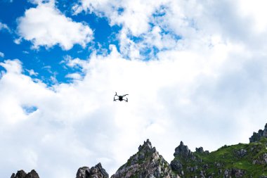 Mavi gökyüzü ve buluta karşı dramatik dağ manzarasında uçan drone silueti. Doğada ticari insansız hava aracı uçuşu