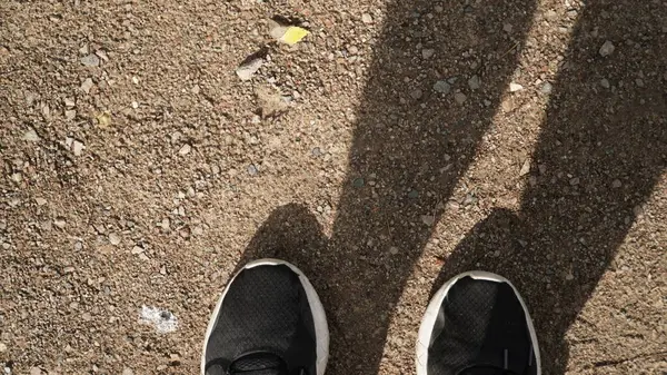 Çakıl üzerinde beyaz tabanlı siyah ayakkabılar fotoğrafçının beyaz tabanlı siyah ayakkabılarını yakalar. Ayakkabının sadece alt yarısı görülebiliyor ve ayakkabı bağcıkları görüş açısında değil. Bu minimalist kompozisyon