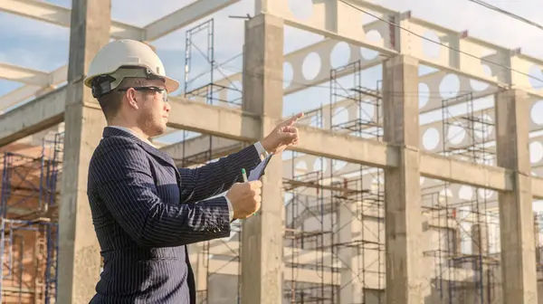 Asyalı yetişkin ustabaşı inşaat projesinin önünde duruyor ve bir inşaat projesini kararlılıkla inceliyor. Mühendis inşaatı denetliyor..