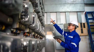 Elektrik bakım mühendisi elektrik sayacını, ana dağıtım panosunu kontrol ediyor ve bir panoya elektrik değerini yazıyor.