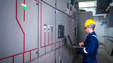 Elektrik mühendisi ana elektrik kontrol panelleri ve anahtarlar, dizüstü bilgisayardaki kayıt değerleri, elektrikçi fabrikadaki elektrik sistemlerini kontrol ediyor..