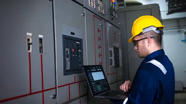 Elektrik mühendisi ana elektrik kontrol panelleri ve anahtarlar, dizüstü bilgisayardaki kayıt değerleri, elektrikçi fabrikadaki elektrik sistemlerini kontrol ediyor..