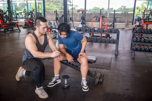 Erkek antrenör, bir fitness merkezinde fiziksel formlarını geliştirmek isteyen bir müşteri için egzersiz programı hazırlıyor.