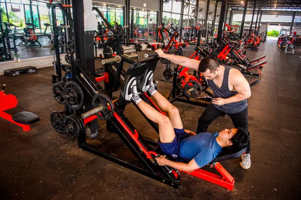 Erkek bir antrenör, makinenin saplarını iterek öğrencisinin bacak pres makinesini kullanmasına yardım ediyor..